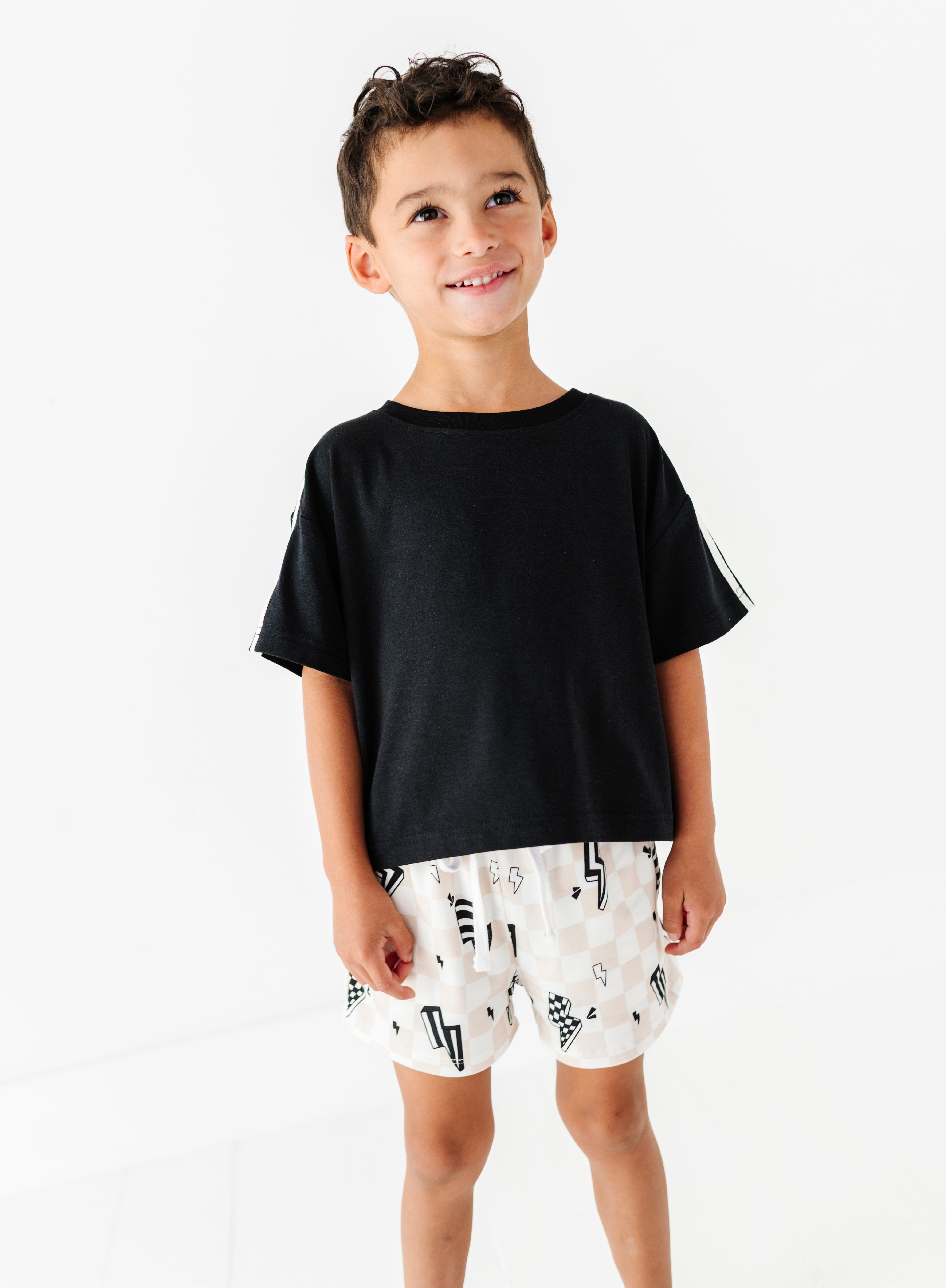 A young boy smiling and looking to the side, wearing a black short-sleeve t-shirt and white shorts with a black graphic print, set against a plain white background.